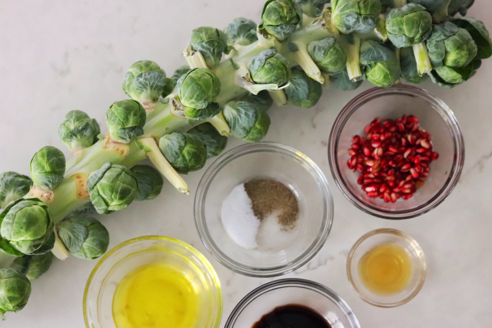 Stalk of Brussels sprouts, a small bowl of pomegranate seeds, olive oil, honey, balsamic vinegar, salt and pepper