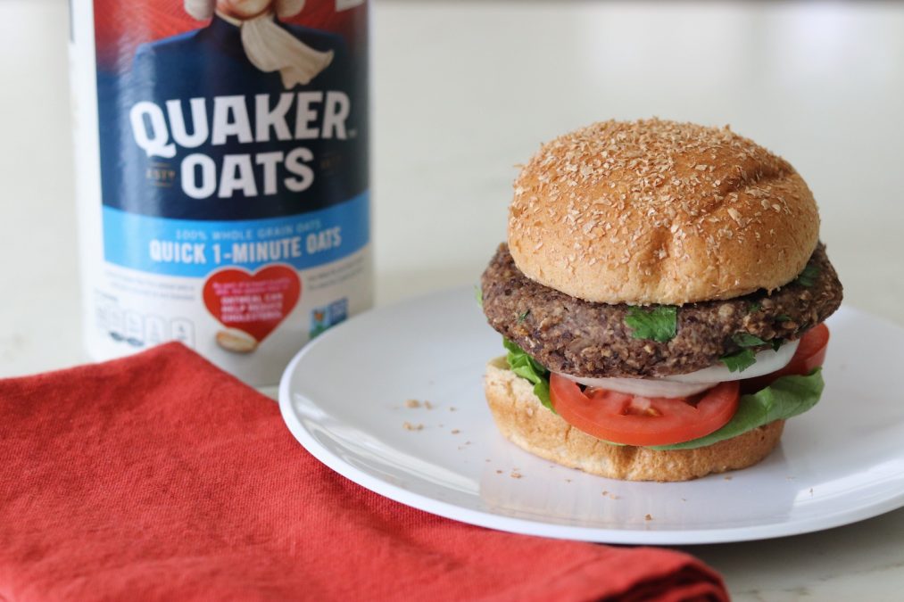 Oat Black Bean Vegetable Burger on a bun with a carton of Quaker Oats in the background