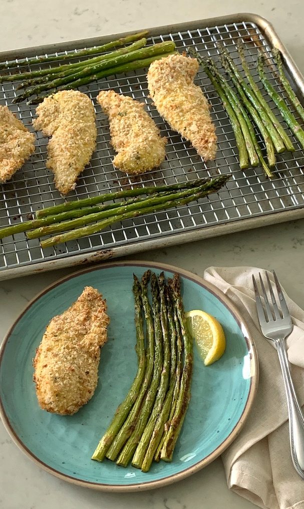 A plate with a portion of crispy parmesan chicken and asparagus and a pan of food in the background