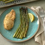 A plate with a portion of crispy parmesan chicken and asparagus and a pan of food in the background