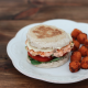 A salmon burger on an English muffin with Lemon-Dijon Aioli, spinach, tomato, and onion on a plate with sweet potato tots
